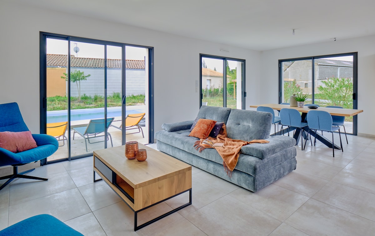 A modern living area is highlighted by large glass doors that offer a view of the pool area. A comfortable gray sofa is paired with a wooden coffee table, while colorful chairs surround a dining table, creating an inviting space for relaxation and meals.