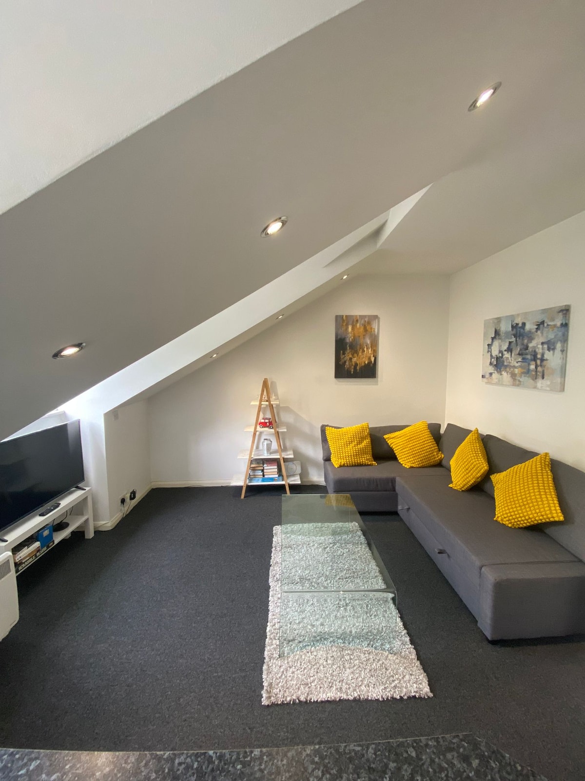A spacious living area features a gray sectional sofa adorned with yellow cushions. A glass coffee table sits on a textured rug, while a ladder-style shelf displays books. Two abstract artworks decorate the walls, and recessed lighting highlights the room's angles.