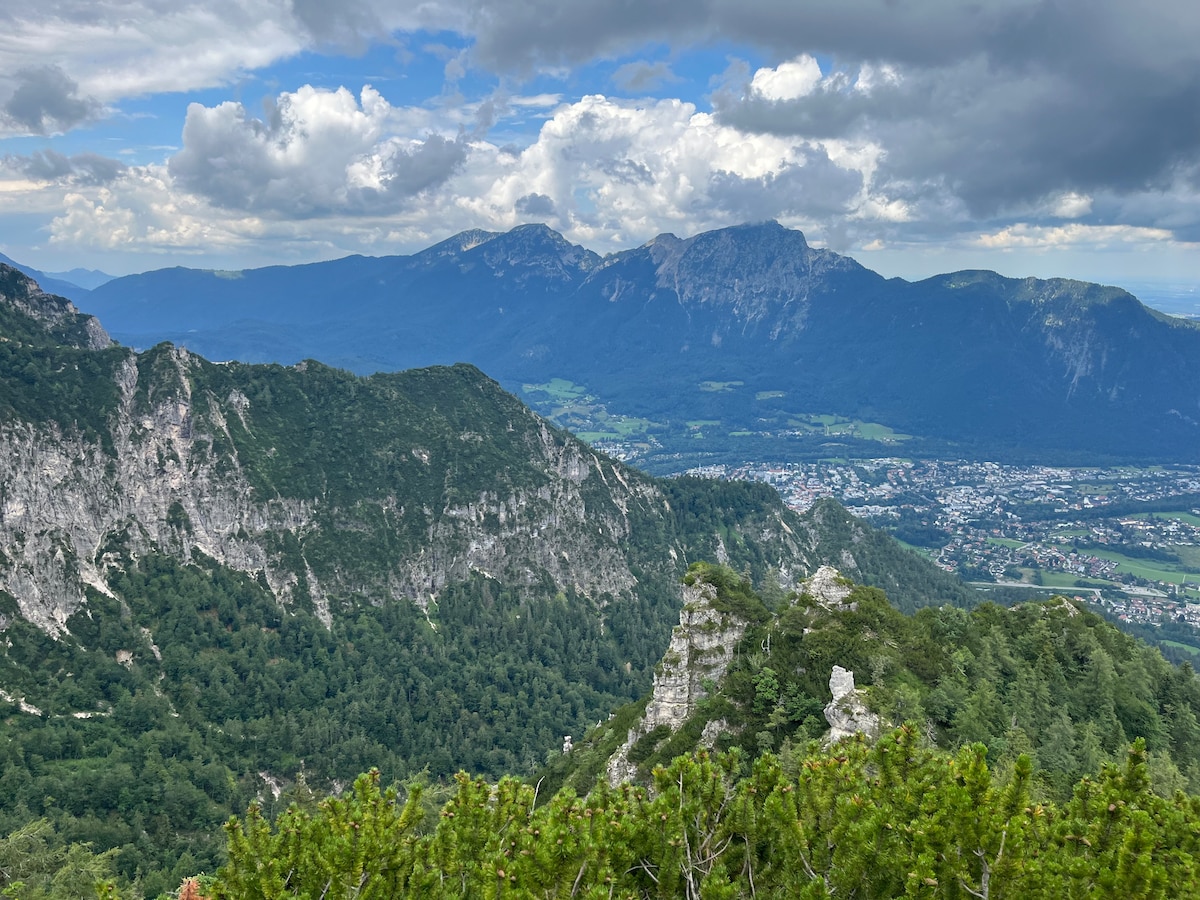 A sweeping mountain view captures rugged peaks and lush valleys under a partly cloudy sky. The greenery of the landscape contrasts with the rocky cliffs, creating a serene and expansive vista that highlights the natural beauty of the region.