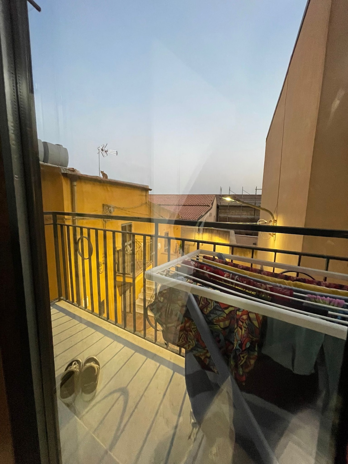 A small balcony is visible, featuring a railing and a modest wooden floor. A clothes rack holds several articles of clothing, while a pair of shoes rests nearby. The view includes neighboring buildings softly illuminated in the evening light.