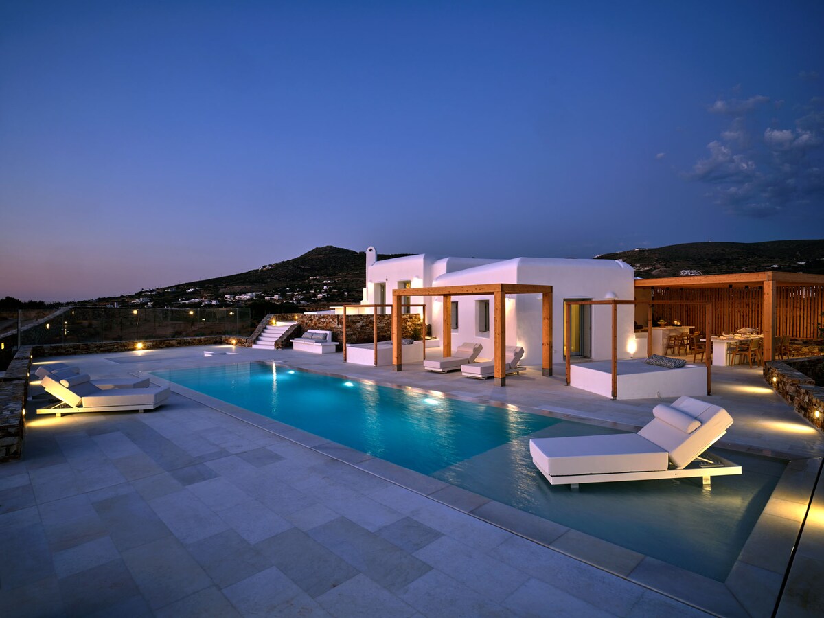 A modern villa is positioned alongside a serene private pool, illuminated subtly against the twilight sky. Multiple sun loungers are arranged beside the pool, offering a relaxing space. The structure showcases wooden accents and expansive outdoor areas, contributing to a tranquil atmosphere.