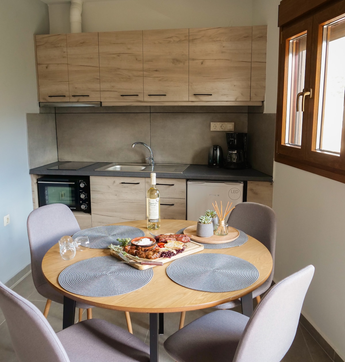 The kitchen area features a modern design with wooden cabinetry and grey countertop. A round dining table is set with two grey chairs, complemented by a platter of snacks and a bottle of wine. Natural light enters through a window, enhancing the space.