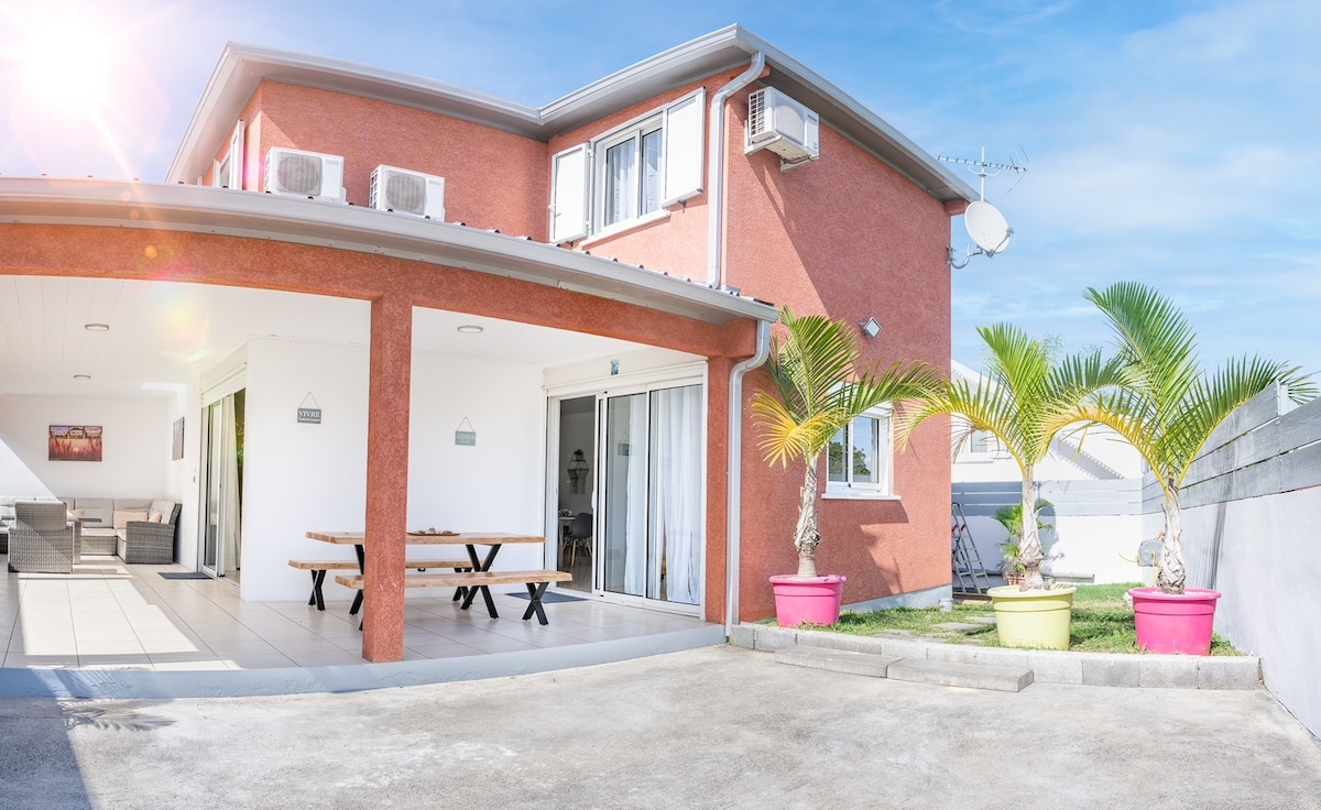 A modern villa exterior features warm red walls and a spacious terrace. Plants in vibrant pink pots add color, while sliding glass doors provide access to the interior. The driveway is paved, and outdoor seating is visible, creating a welcoming entryway for guests.