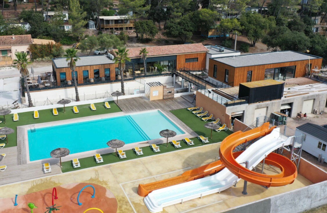 An aerial view of a communal area featuring a large swimming pool surrounded by sun loungers. An orange water slide is positioned near a sandy play area with colorful equipment for children. Lush greenery and buildings with wooden facades are visible in the background.