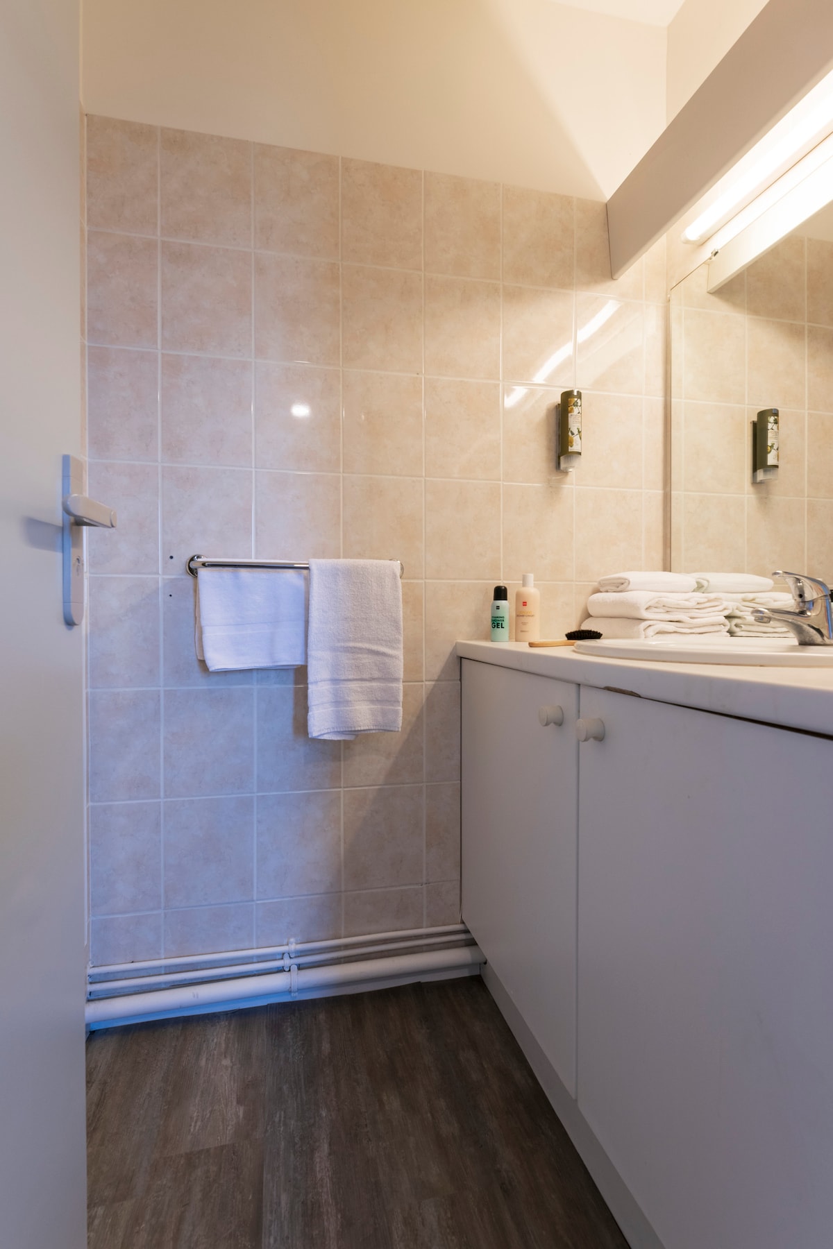 A compact bathroom features tiled walls with a clean aesthetic. It includes a modern sink area, a large mirror with overhead lighting, and a towel rack displaying white towels. Storage cabinets beneath the sink provide additional space.