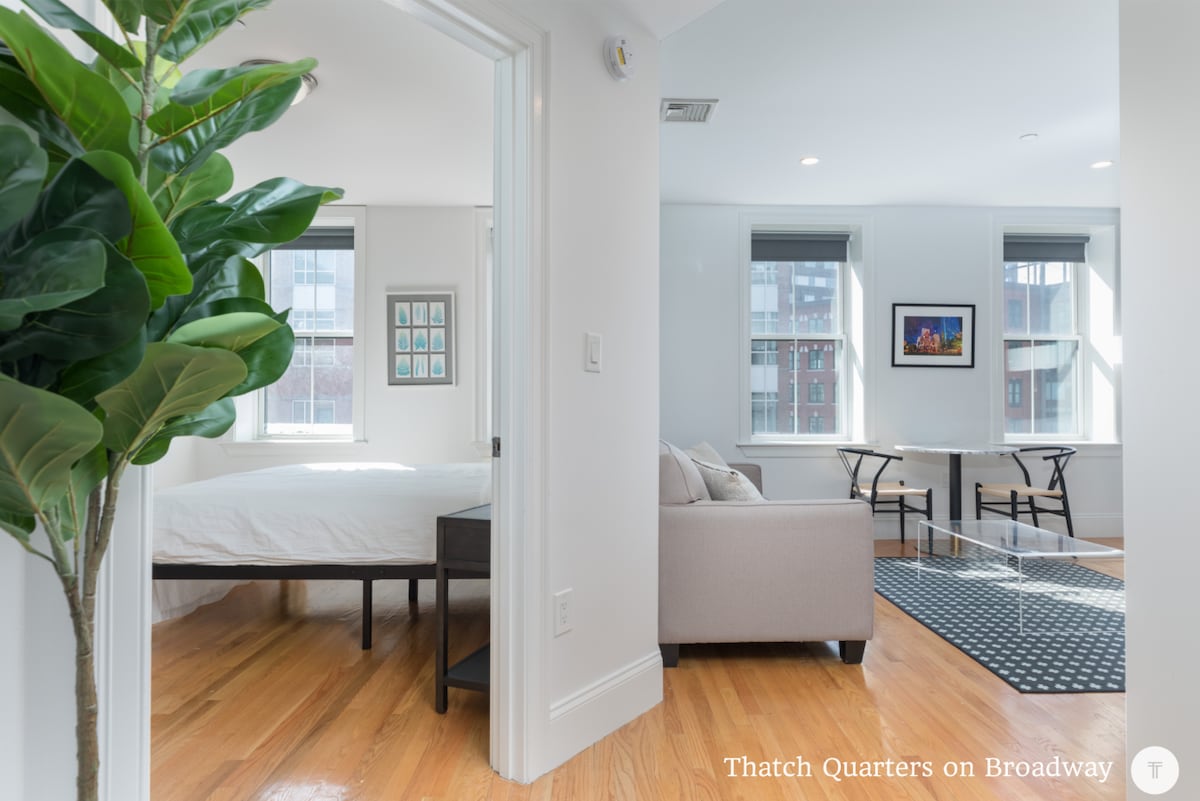 A bright and airy room is presented, showcasing a spacious bed framed by two large windows that allow ample natural light. A seating area with a couch and small dining table can be seen to the right, complemented by polished hardwood floors and a touch of greenery in the foreground.