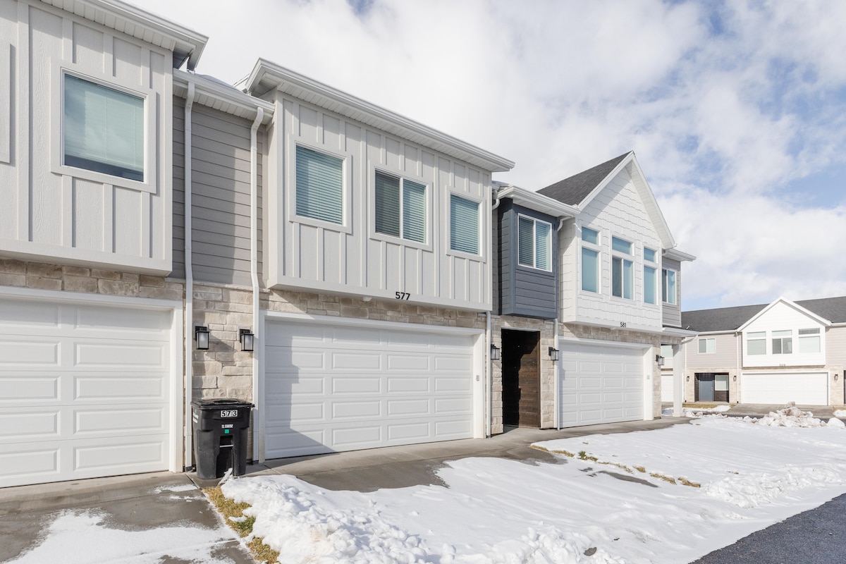 A modern townhouse exterior is displayed, featuring a combination of light gray and white siding. The image shows three garage doors, with two on the left and one larger door on the right. Snow is visible on the driveway and ground, indicating winter conditions.