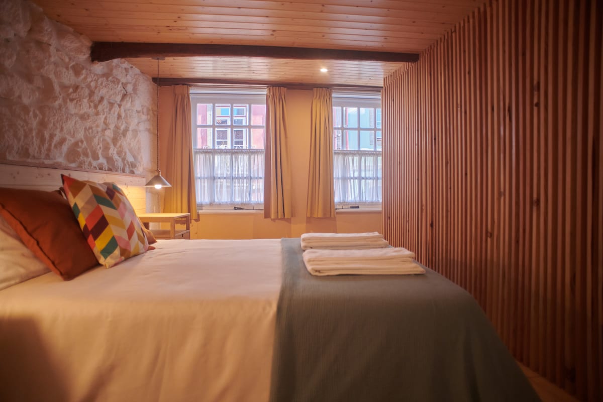 A cozy room features a spacious bed with neutral and patterned pillows. Two windows allow natural light to illuminate the space, complemented by warm wood paneling and soft curtains. A small bedside lamp and wooden table provide functional decor.