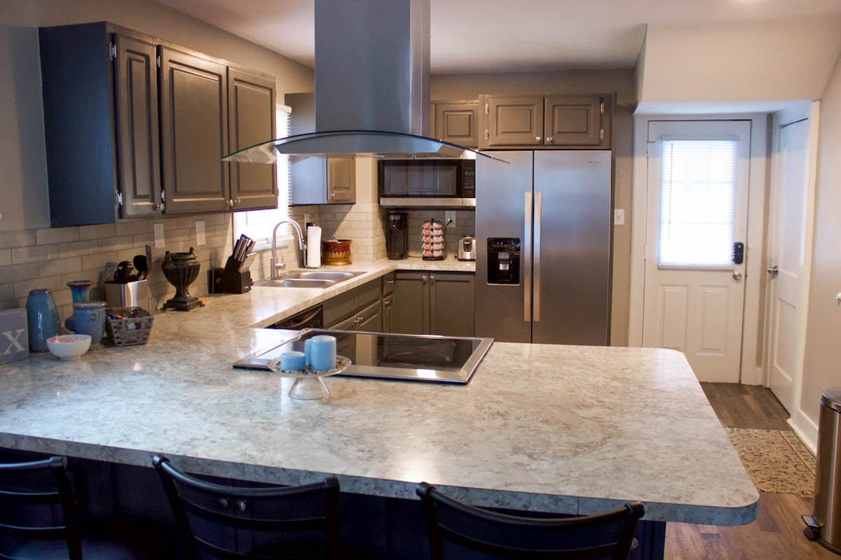 The kitchen features modern cabinetry and a large countertop bar with seating for four. Stainless steel appliances, including a refrigerator and oven, are visible. Natural light streams in through the window, illuminating the spacious layout, which includes a stovetop and sink area.
