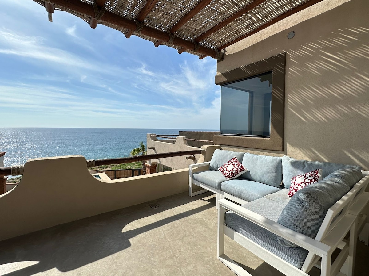 A spacious outdoor seating area features a comfortable sectional sofa with light blue cushions and decorative red pillows. The scene includes an expansive view of the ocean under a partly cloudy sky, with natural light streaming in through large windows.