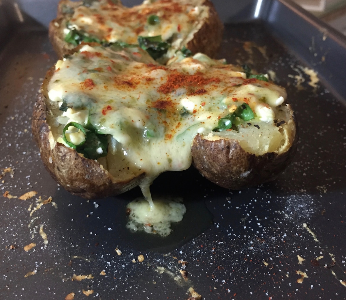 Two baked potatoes sit on a dark baking tray, topped with melted cheese, spinach, and a sprinkle of paprika. The potatoes appear fluffy inside, with a golden, creamy layer of cheese pooling slightly over the edges.