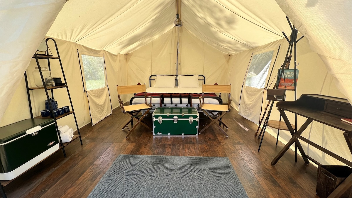 The interior of the tent showcases a spacious layout with a king-sized bed framed by wooden supports. A green cooler and a decorative trunk serve as functional storage. Indoor seating is available with a stylish table, while side shelves offer additional convenience.