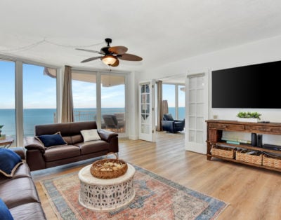 A spacious living area features a comfortable couch, a decorative coffee table, and large windows that provide a view of the ocean. Natural light floods the room, highlighting the airy atmosphere. A television is mounted on the wall, and a decorative rug adds warmth to the space.