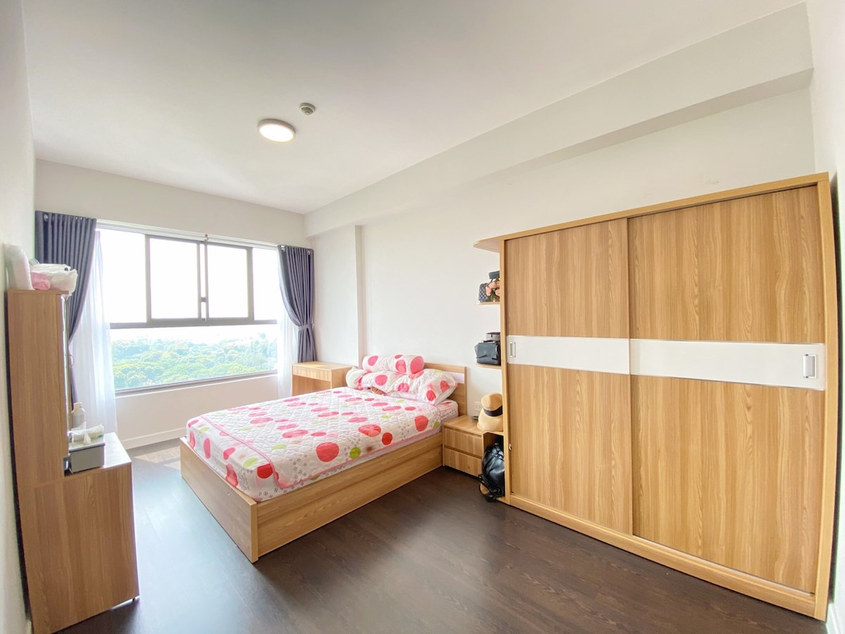 A well-lit bedroom features a spacious bed with a patterned pink and white duvet. A large window offers scenic views, while a wooden wardrobe and bedside table provide ample storage. Dark flooring contrasts with the bright walls and soft curtains, enhancing the room's comfort.