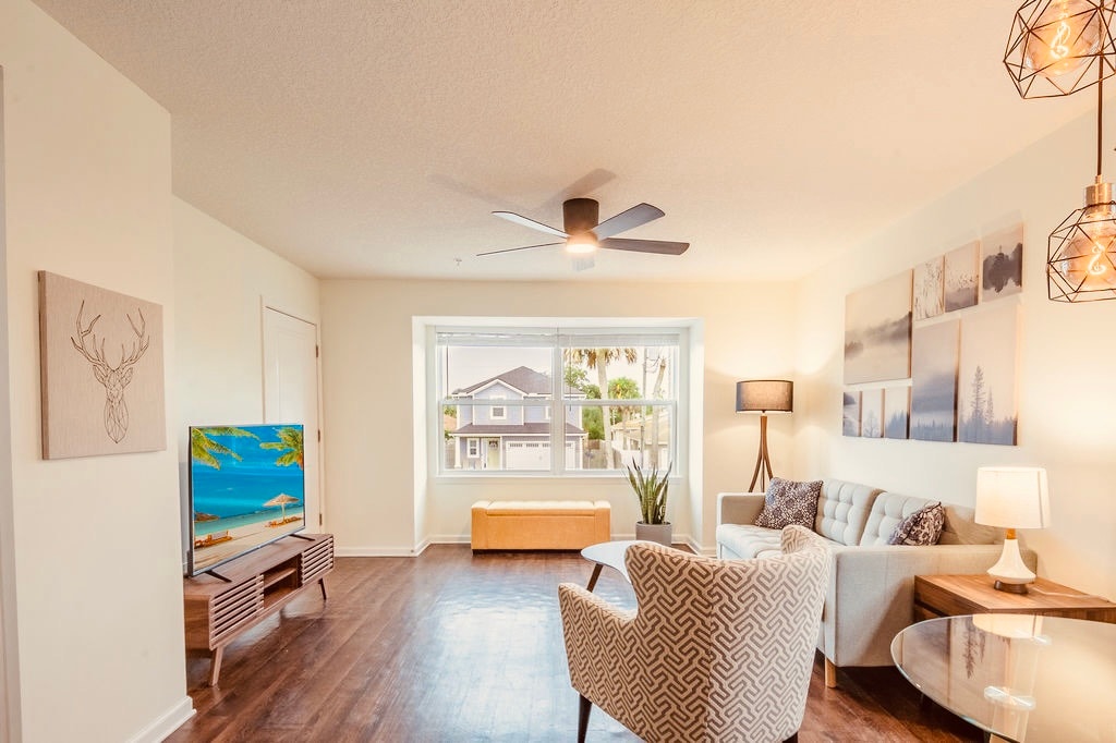 The bright living area features a large window allowing natural light to fill the space. A comfortable seating arrangement includes a patterned chair and a cozy couch. A mounted TV and a round dining table enhance the functionality, while a ceiling fan adds a touch of modern decor.