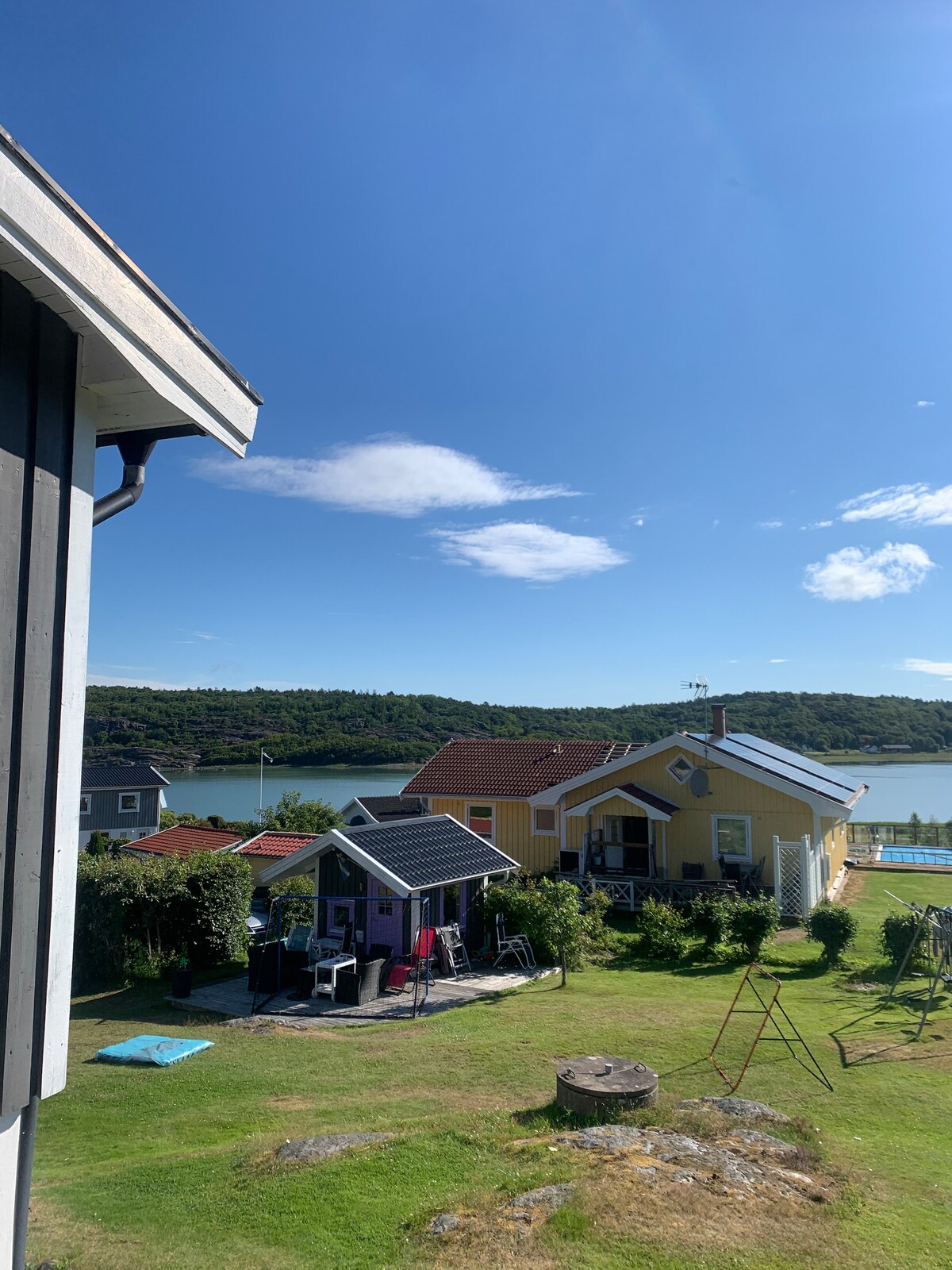 A serene view reveals a cluster of charming houses with vibrant colors set against lush greenery and a calm water backdrop. The sky is clear, showcasing soft clouds, while a grassy area invites relaxation and leisure activities.