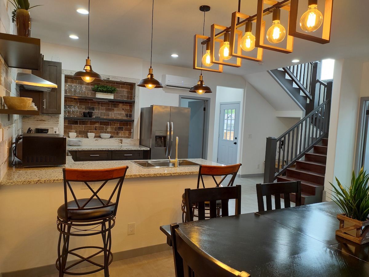 An open-concept kitchen and dining area is highlighted by modern pendant lighting above the bar. The kitchen features granite countertops and tasteful cabinetry, while a dining table is set with dark wooden chairs. A staircase is visible in the background, leading to the upper level.
