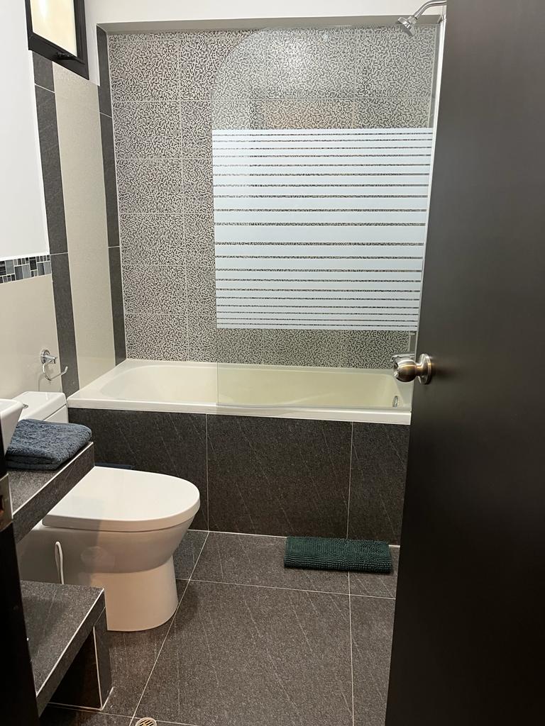 A modern ensuite bathroom is showcased, featuring a bathtub and a toilet. The walls are adorned with a textured pattern, while a sliding glass panel provides separation for the shower area. Dark tiles create a sleek look, complemented by a small rug on the floor.