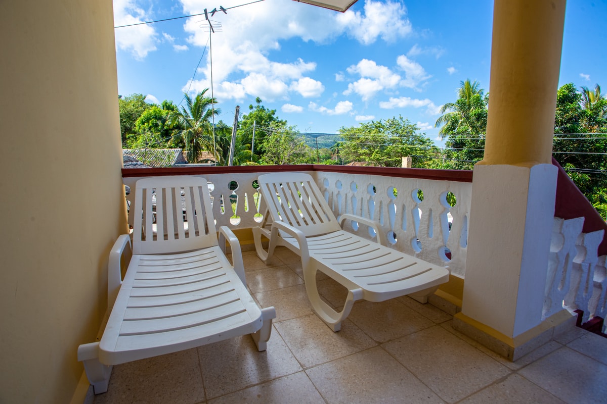 Two sun loungers are positioned on a sheltered terrace, offering views of lush greenery and a blue sky. The terrace features white railing patterns, enhancing the outdoor space and inviting relaxation in a tranquil setting.