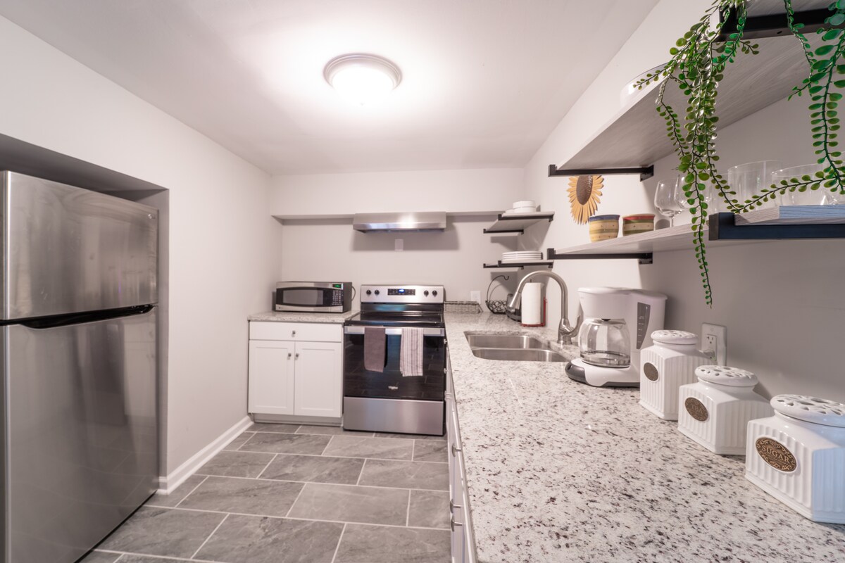 A compact kitchen area is equipped with modern stainless steel appliances, including a refrigerator, microwave, and stove. Shelving holds kitchen essentials, while a sleek countertop offers ample preparation space. Neutral-colored cabinets blend with the granite-like surface, creating a clean and functional cooking environment.
