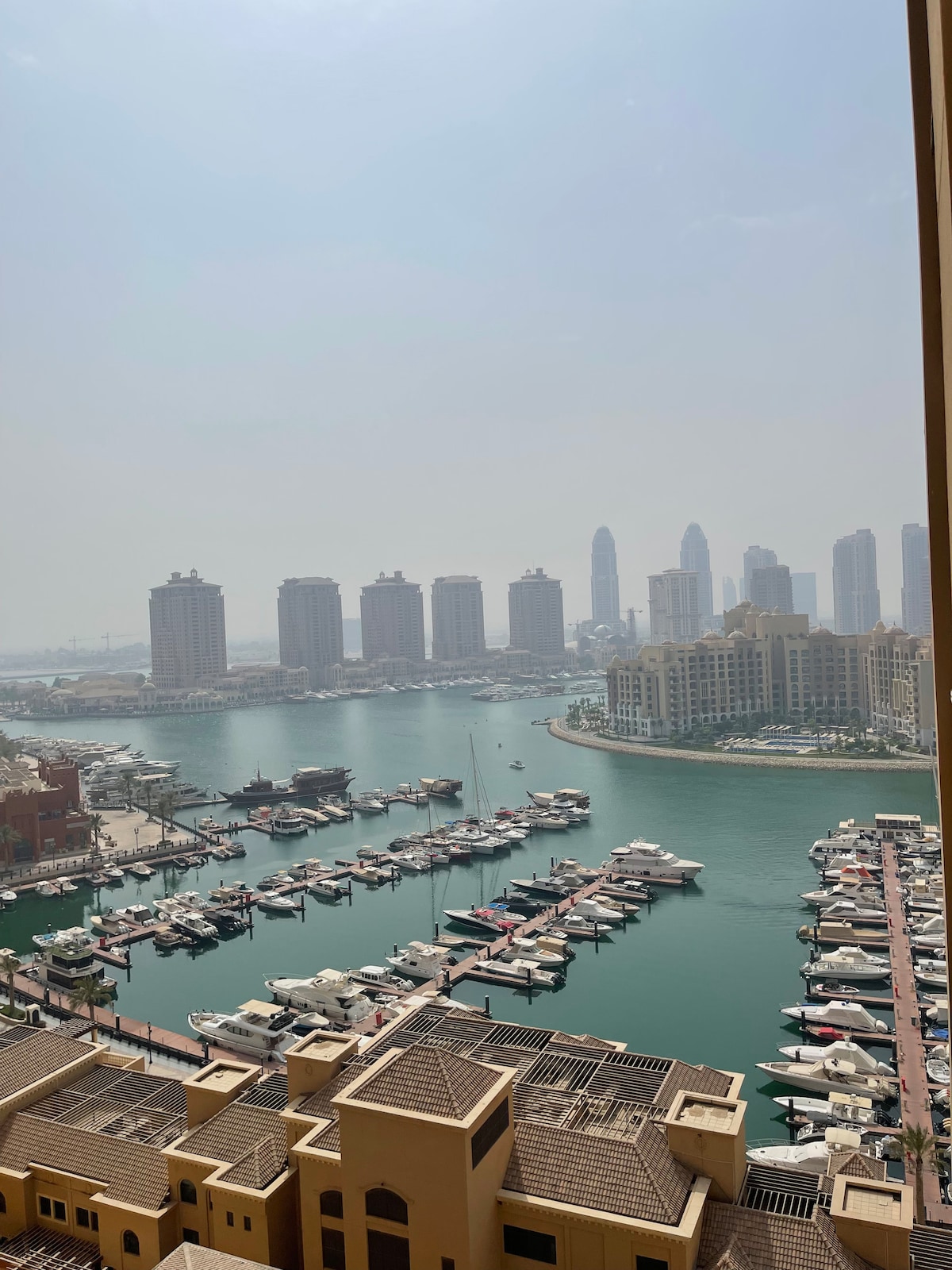 A scenic view of a marina is visible, showcasing numerous boats anchored along the water. Towering buildings rise in the background, while the gentle curves of the marina's shoreline can be seen. A soft haze adds a serene quality to the skyline.