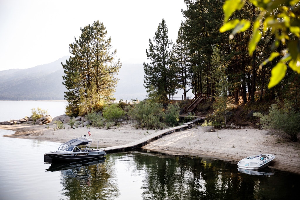 A serene lakeside view is presented, featuring a wooden dock leading to calm waters. Two boats are anchored nearby, surrounded by lush trees and gentle hills in the background. Soft sunlight highlights the sandy beach, creating a warm and inviting atmosphere.
