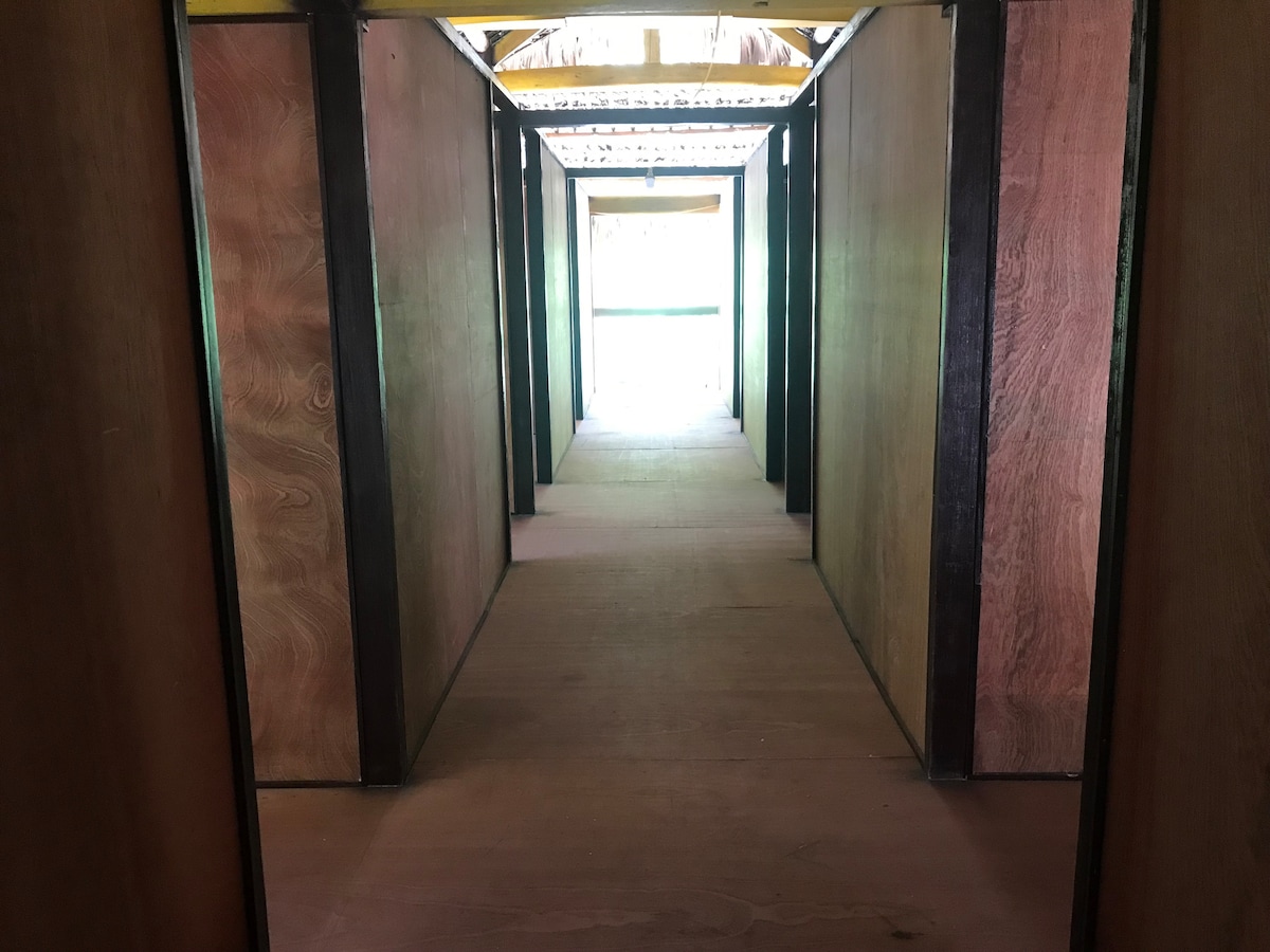 A long corridor with wooden walls leads towards an open area at the far end. Natural light filters in, enhancing the warm tones of the wood. The floor is made of smooth timber, creating a clean and inviting path.