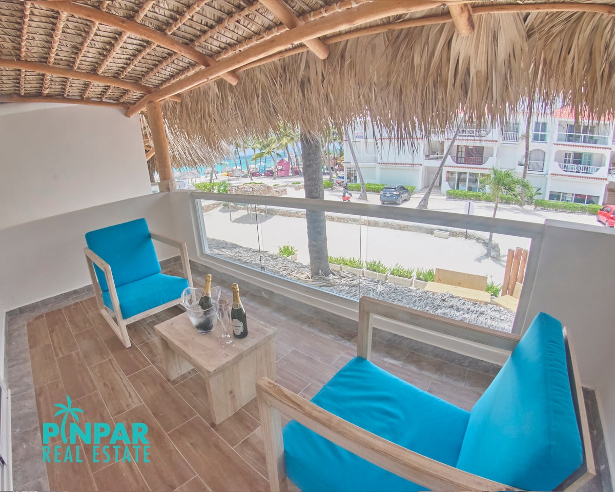 A private balcony features two blue chairs and a compact wooden table, complementing the thatched roof overhead. A bottle of champagne is placed on the table, with a view of the beach and palm trees visible through the glass railing.