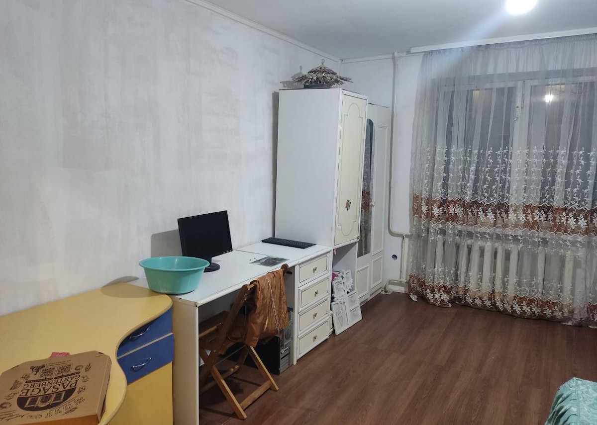 A functional workspace is present, featuring a desk with a computer and a bucket. A wardrobe offers storage options, while a window adorned with light curtains allows natural light into the room. The warm wood flooring complements the neutral walls.