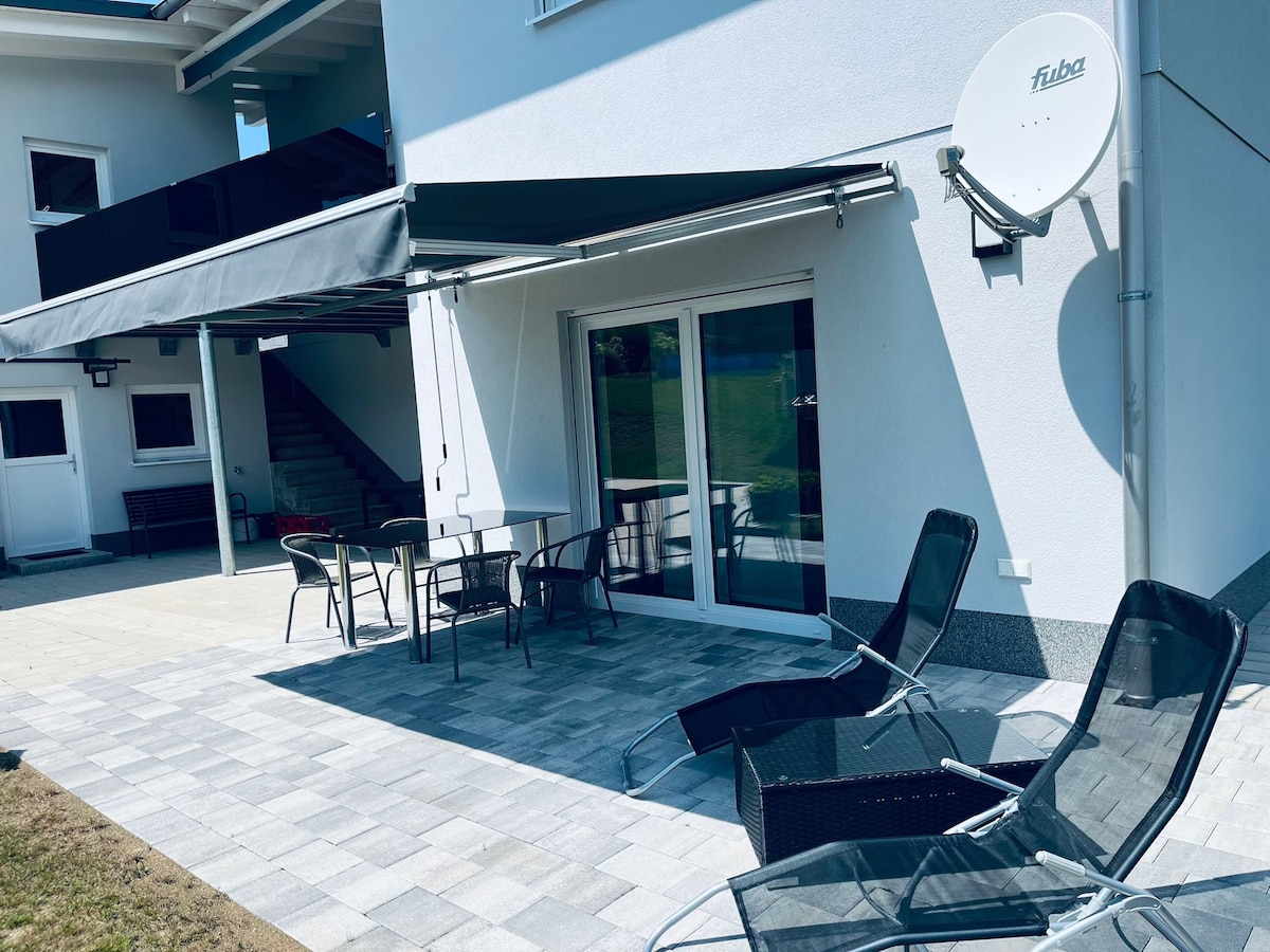 A covered outdoor seating area is showcased, featuring a small table and four chairs, alongside two lounge chairs. An awning provides shade, and sliding glass doors lead into the living space. A satellite dish is mounted on the exterior wall, completing the inviting patio setup.