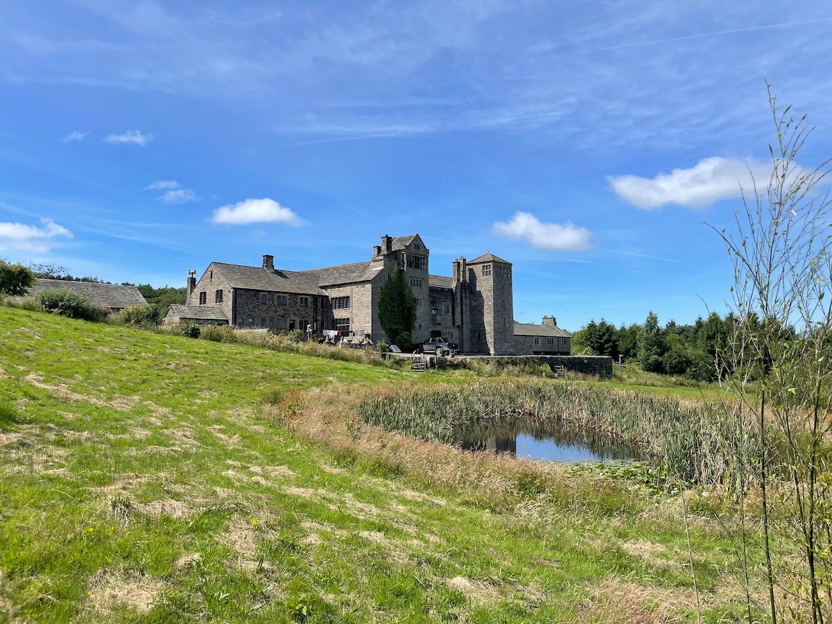 A historic stone building sits gracefully on a hilltop, surrounded by lush greenery and a tranquil body of water. The structure features various architectural elements, including towers and multiple windows, under a bright blue sky accentuated by scattered clouds.