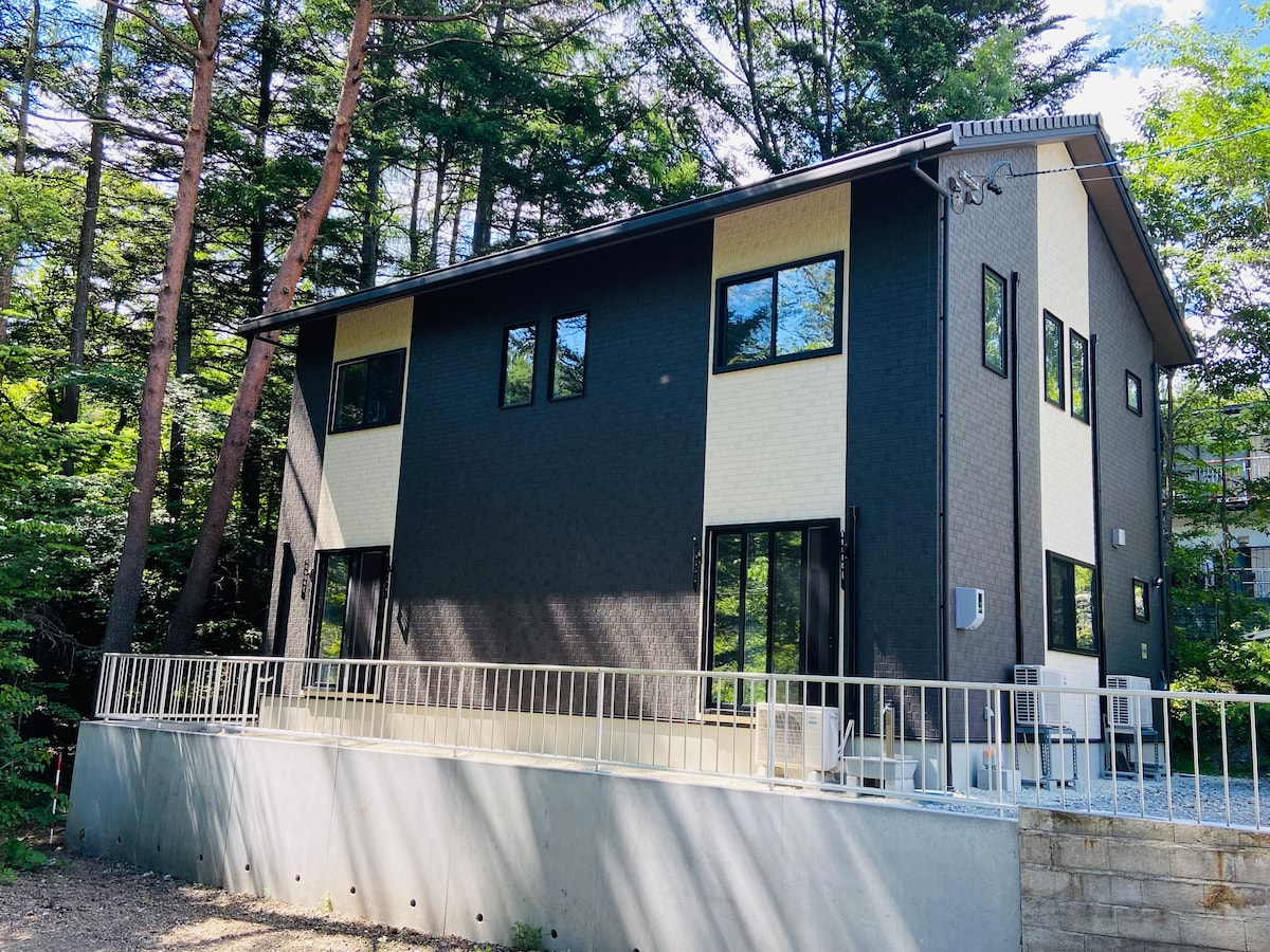 A modern two-story building features a combination of black and light-colored siding. Large windows allow for natural light, surrounded by a lush green landscape. A simple railing encloses a gravel area at the base, complementing the clean lines of the architecture.