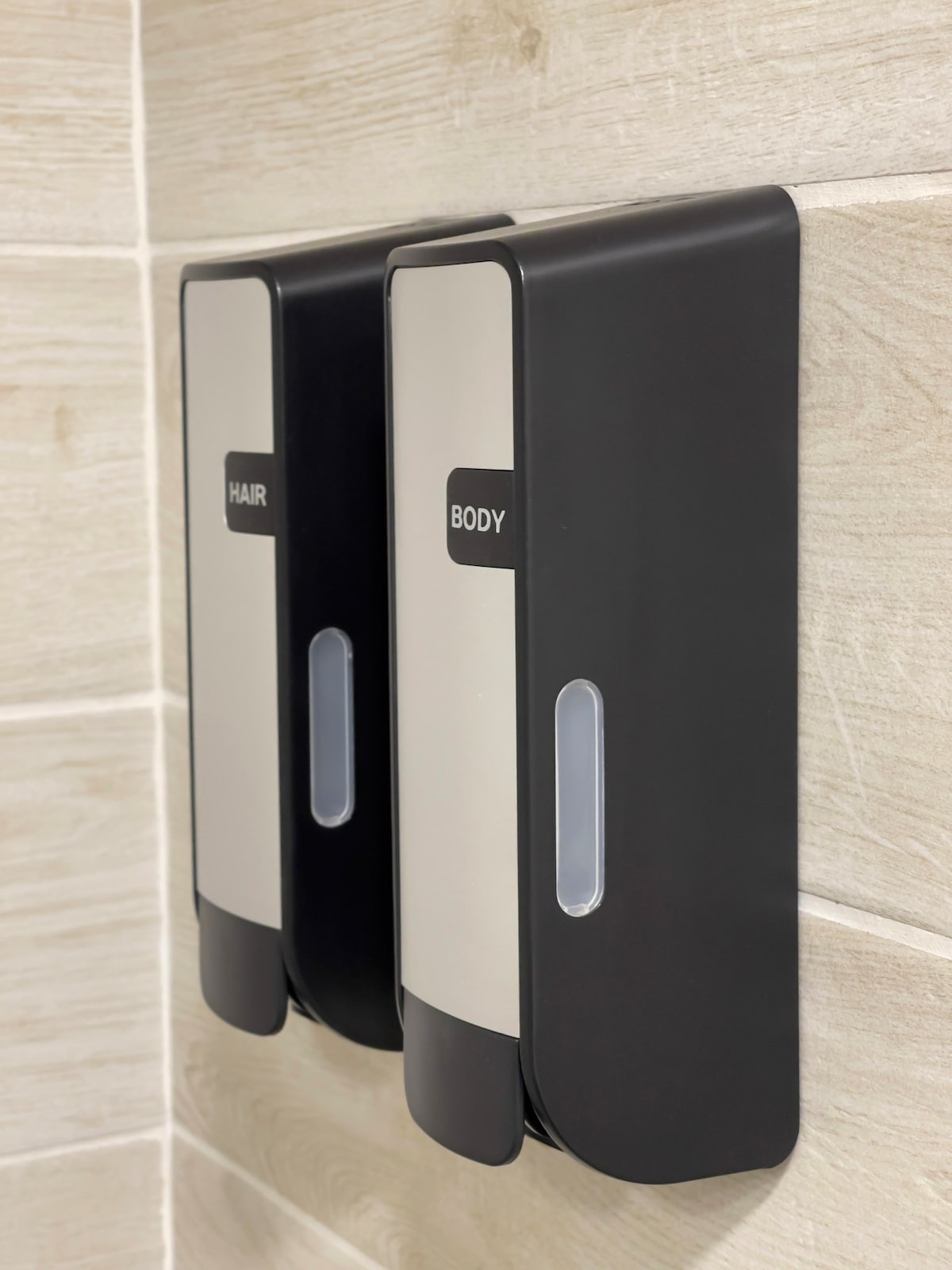 Two wall-mounted dispensers are visible, labeled for hair and body products. The sleek, black and silver design adds a modern touch to the bathroom environment. The dispensers feature transparent sections for easy monitoring of product levels.