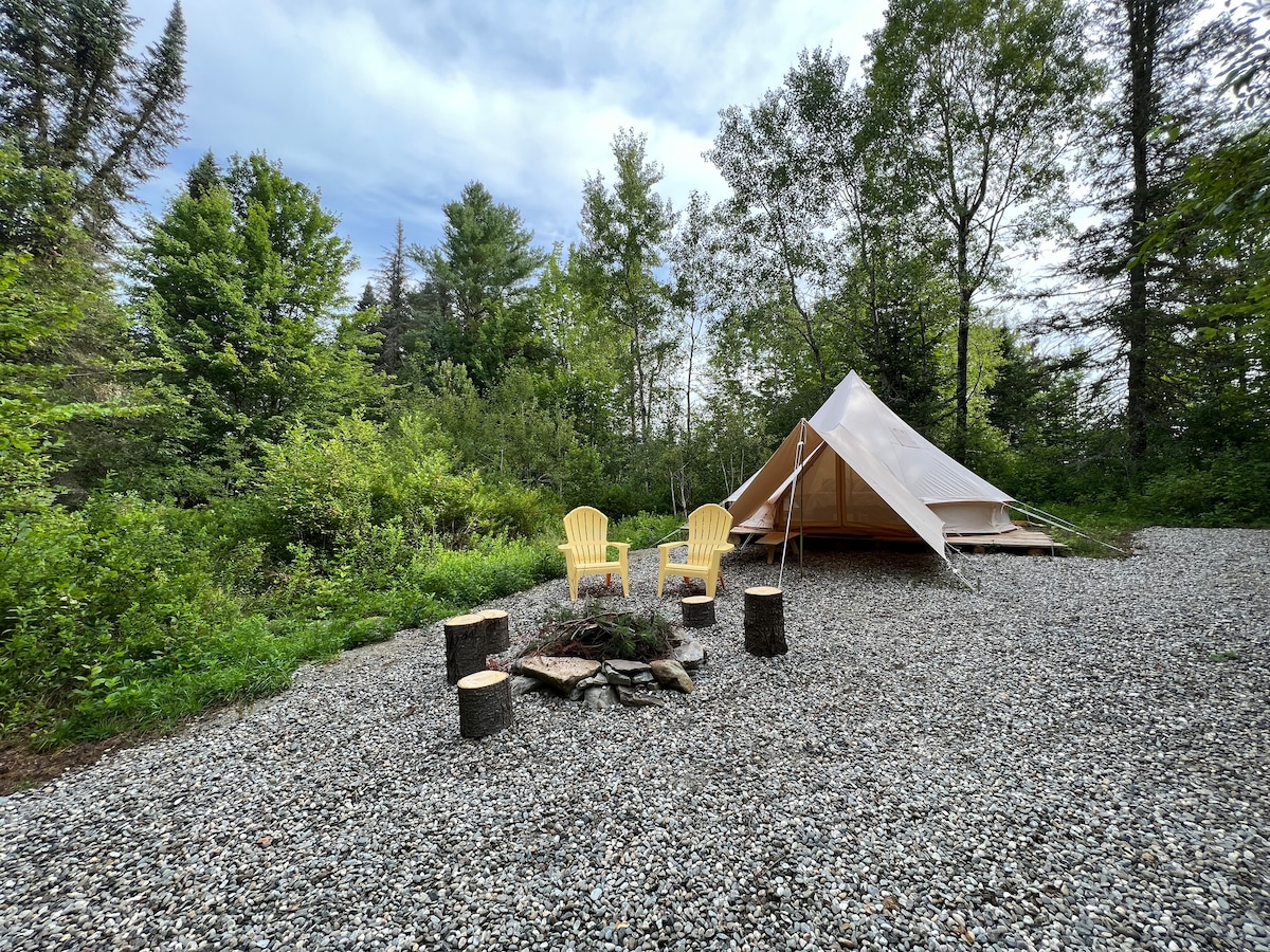 A spacious canvas bell tent is nestled among lush greenery, set upon a gravel area. Two yellow Adirondack chairs frame a small fire pit made of logs and stones, surrounded by vibrant foliage and trees under a serene sky.