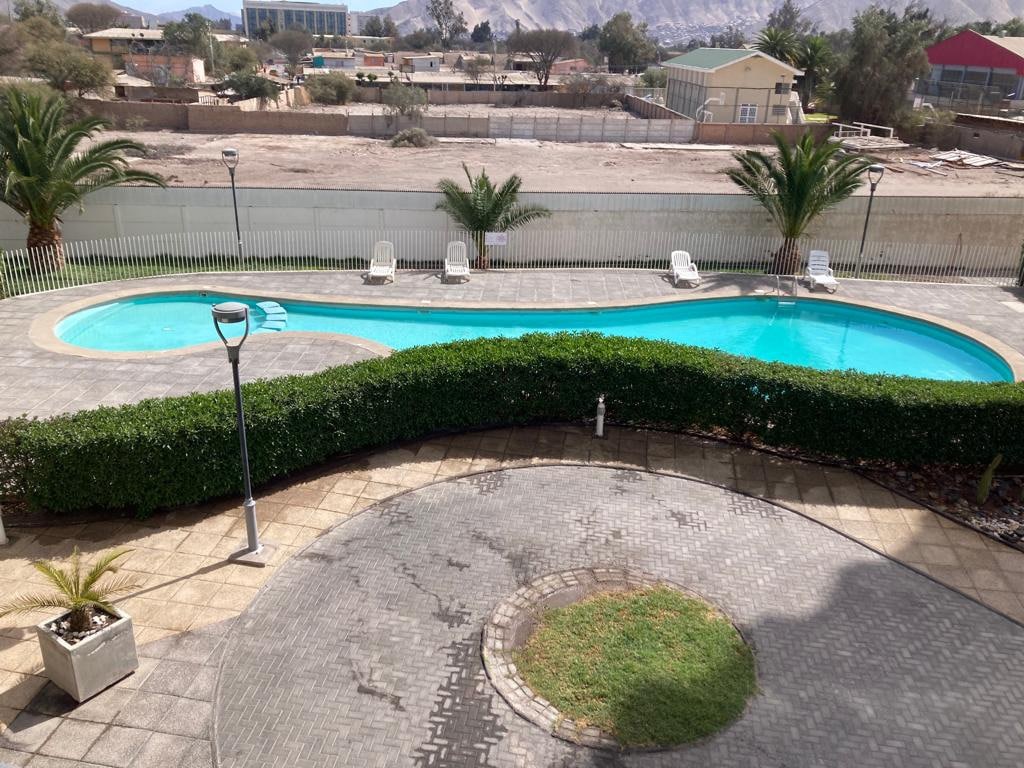 A serene outdoor pool area features a curved pool surrounded by lush green hedges. Several white lounge chairs are placed beside the pool, and a well-maintained pathway is visible. The scene is complemented by palm trees in the background, enhancing the tranquil environment.
