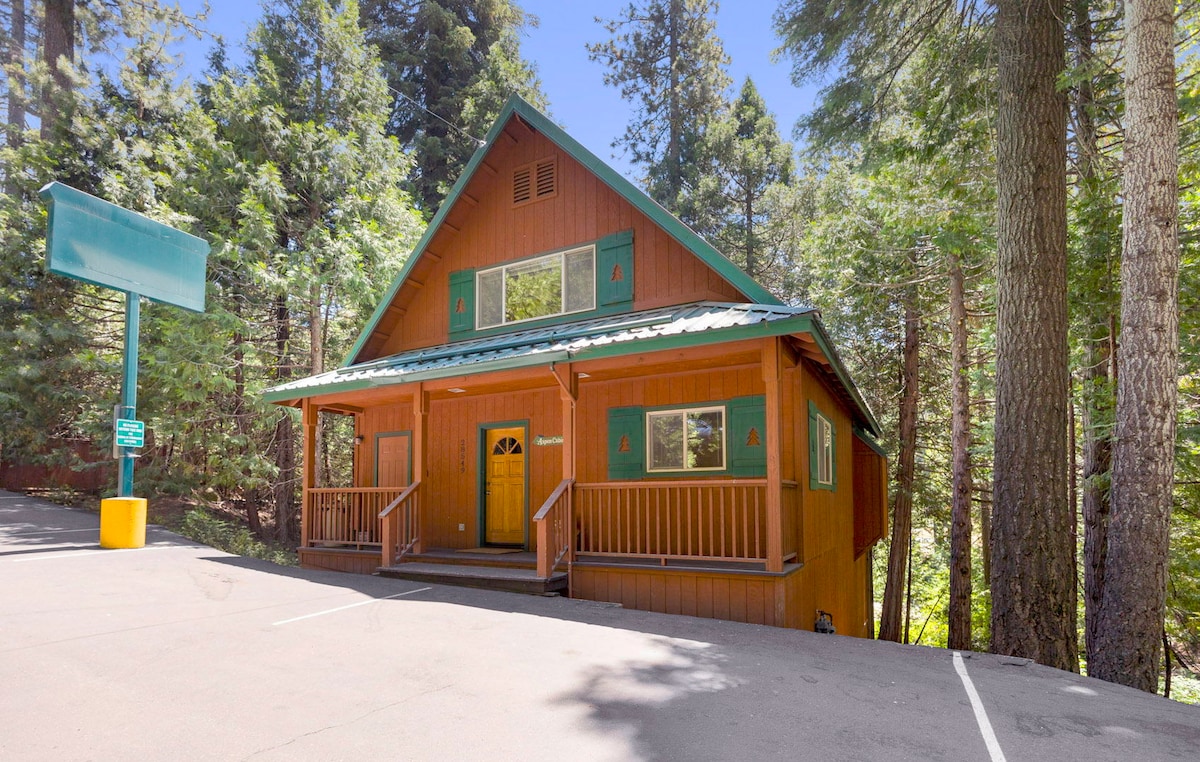 A spacious cabin exterior is surrounded by tall trees, featuring a red wood façade and a sloped green roof. A welcoming front porch with wooden railings leads to the main entrance, while a paved driveway accommodates parking.