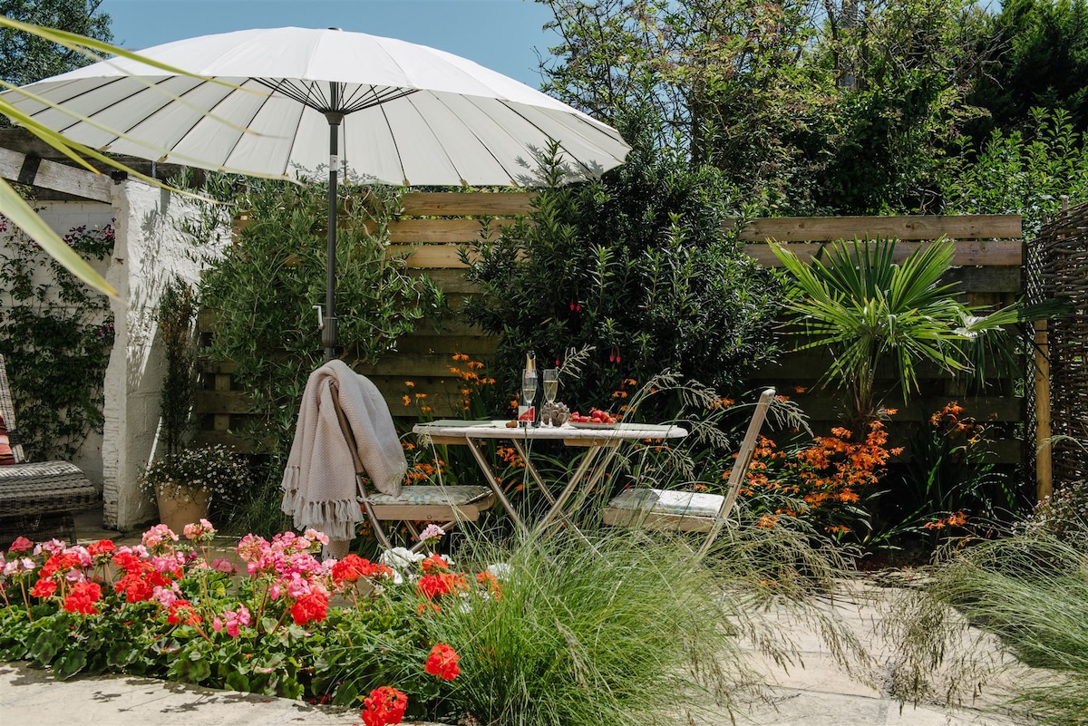 A private patio area features a round table with two chairs, sheltered by a large umbrella. Bright flowers, including geraniums, frame the space, surrounded by lush greenery. A cozy throw is draped over one chair, adding a subtle touch to the inviting outdoor setting.