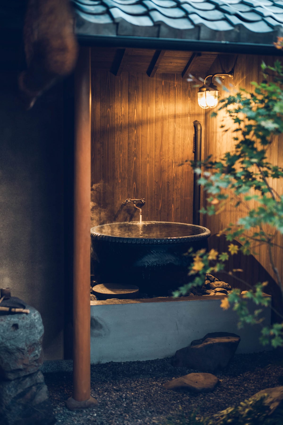 A half-outdoor bath crafted from dynamic pottery is situated under a wooden roof, with steam rising gently. Soft light illuminates the area, highlighting the natural textures of the surrounding wooden walls and stone details.