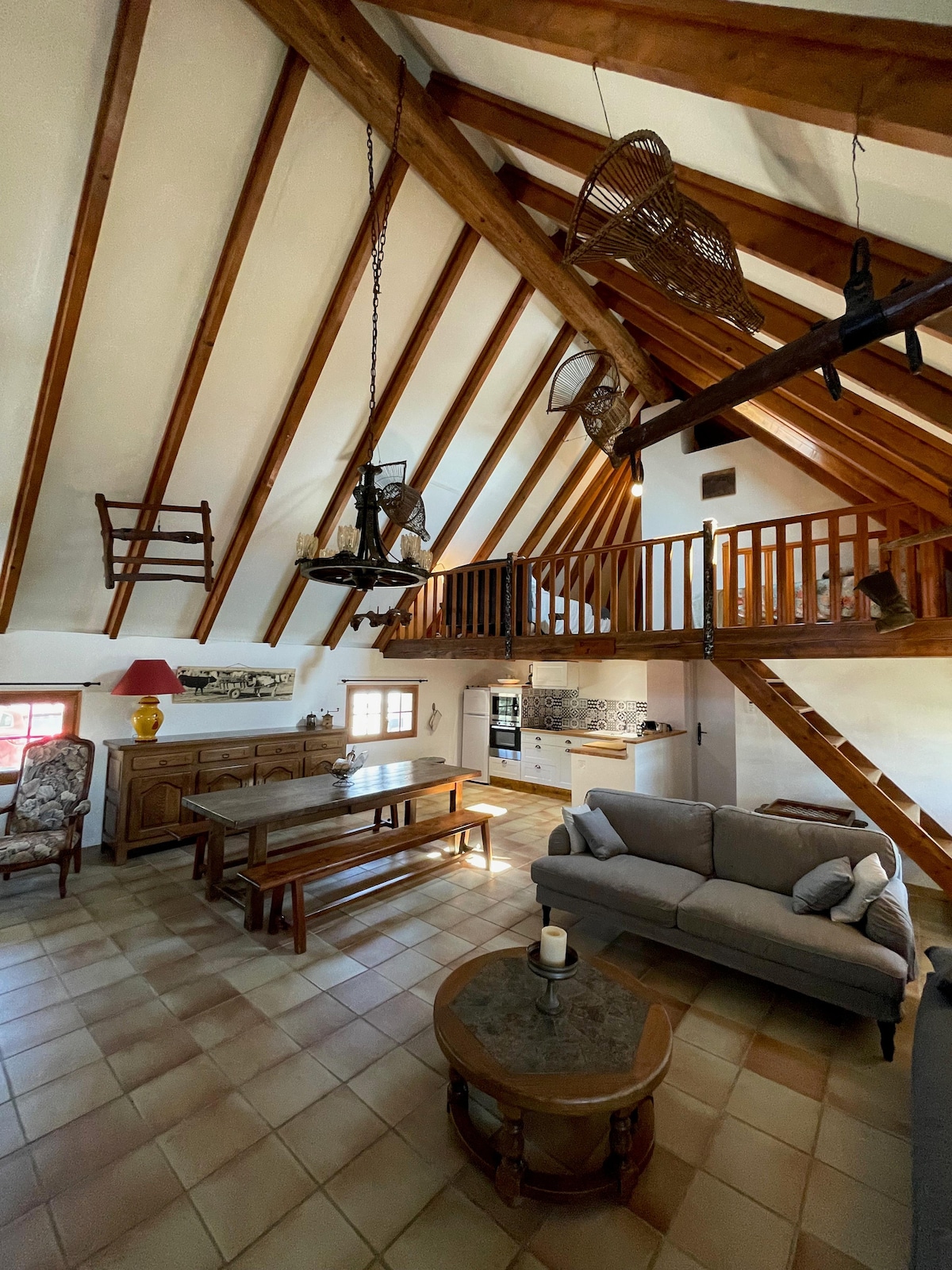 The living space features a high, wooden beam ceiling with a rustic charm. A spacious wooden dining table is placed at the center, accompanied by a comfortable sofa. A well-equipped kitchen is visible in the background, enhancing the open layout of the area.