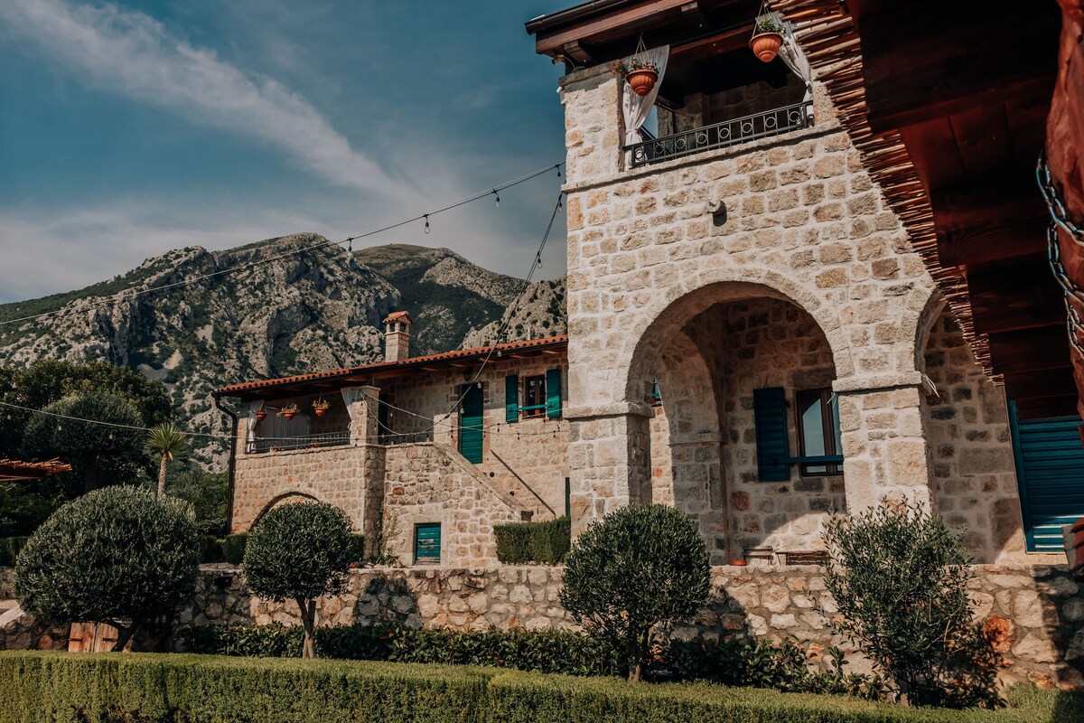 A stone villa is framed by olive trees and manicured greenery, showcasing its rustic architecture. The building features arched doorways and windows, with mountain peaks visible in the background. String lights add a welcoming touch to the outdoor ambiance.