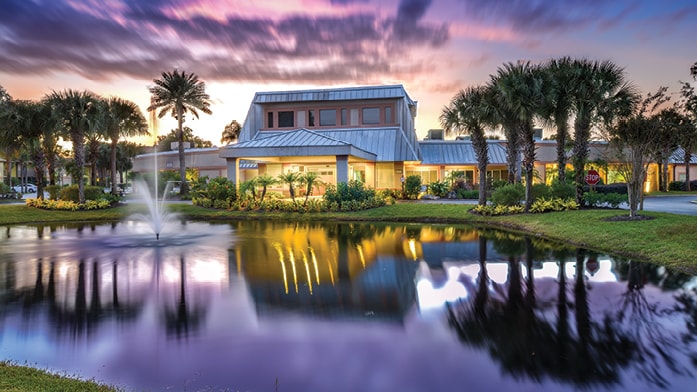 A tranquil pond reflects the beautifully lit exterior of the resort, surrounded by lush landscaping and palm trees. The sky displays soft hues of purple and orange as dusk approaches, creating a serene atmosphere.