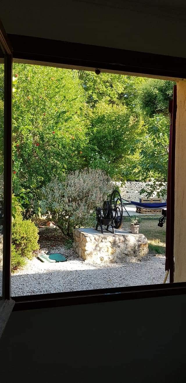 A view from a window showcases a landscaped garden featuring lush greenery and decorative stone elements. A vintage-style wheel is positioned on a stone platform, while a hammock can be seen in the background, inviting moments of relaxation.