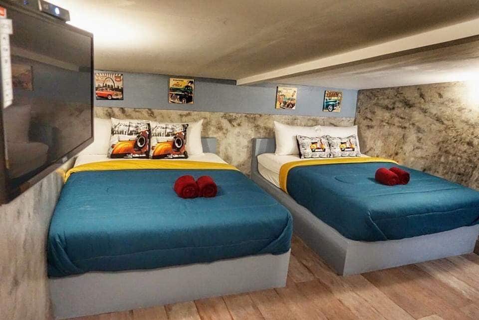 A well-appointed bedroom features two spacious beds dressed in blue covers, complemented by yellow accents. Decorative wall art enhances the space, and a television is mounted on the wall opposite the beds. Warm lighting creates an inviting ambiance throughout the room.