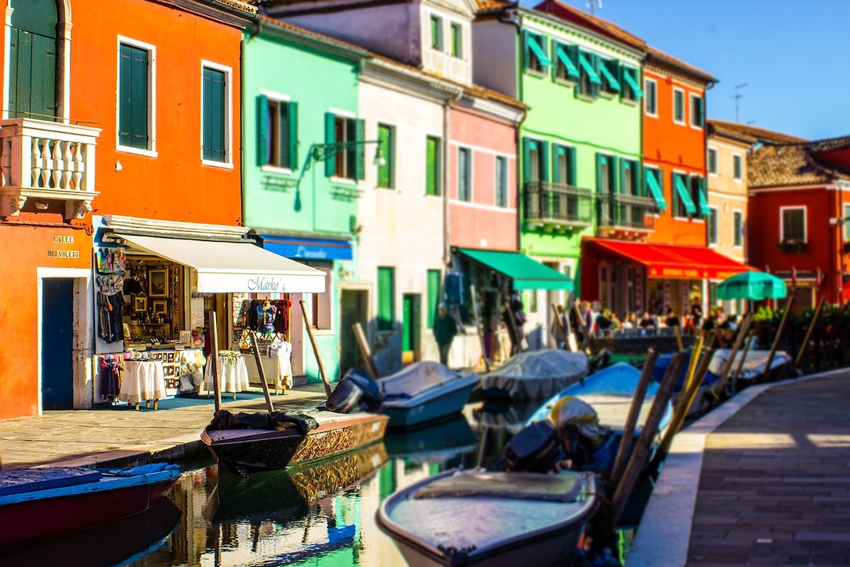 A vibrant street scene in Burano features colorful buildings lining a canal. Small boats are moored along the water's edge, and a shop displays various goods beneath a white awning. Bright hues of orange, green, and blue characterize the architecture.