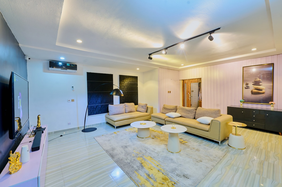 A spacious living area is featured, with a large sectional sofa arranged around white coffee tables. Soft lighting highlights a minimalist design, complemented by a decorative rug. A large television is mounted on the wall, and a modern floor lamp stands beside the sofa.
