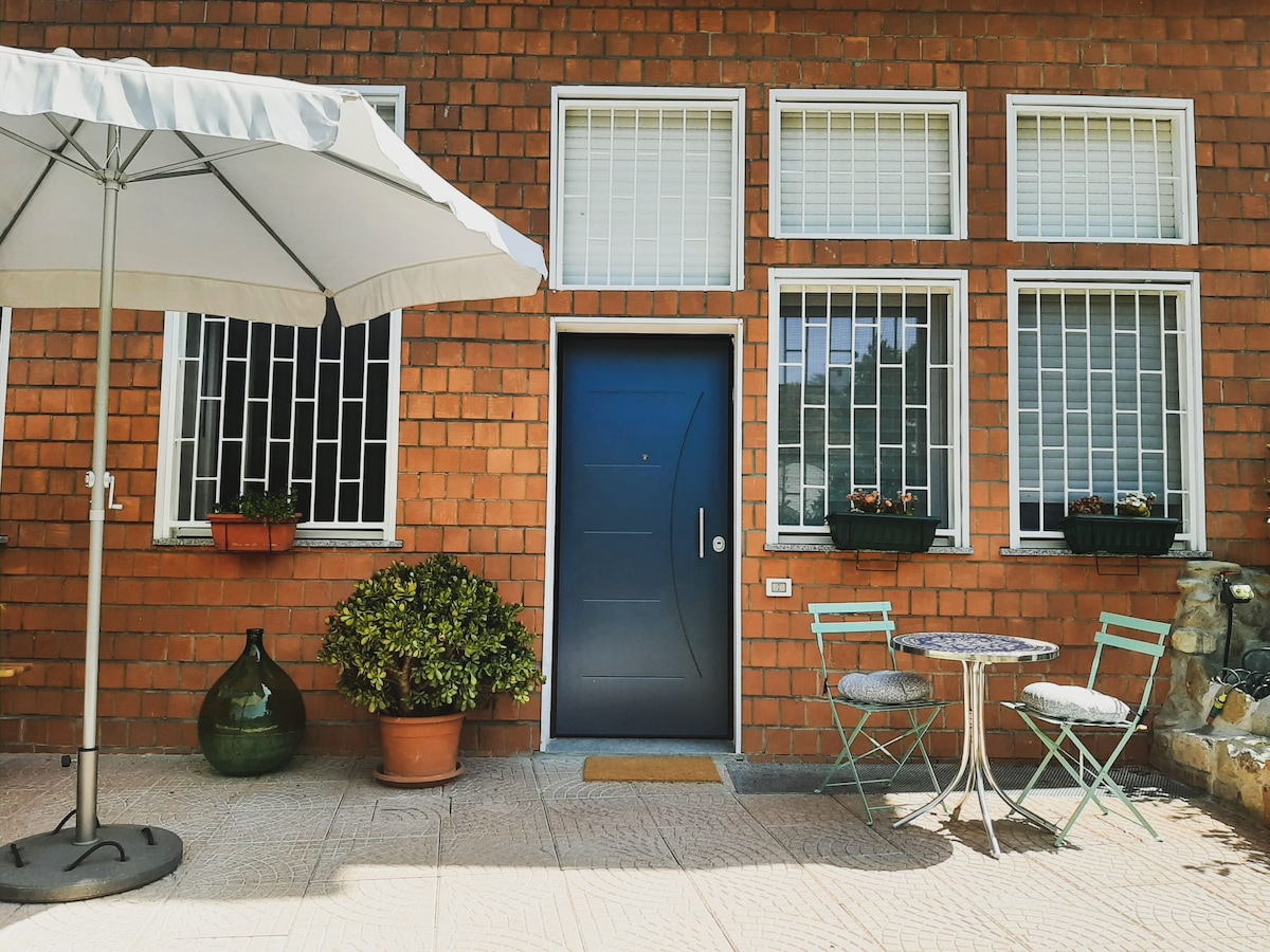 An entrance to the apartment features a modern door framed by large, grid-patterned windows. A small patio is adorned with a round table and two teal chairs. Potted plants and a decorative glass bottle add to the welcoming outdoor space, complemented by a large parasol overhead.