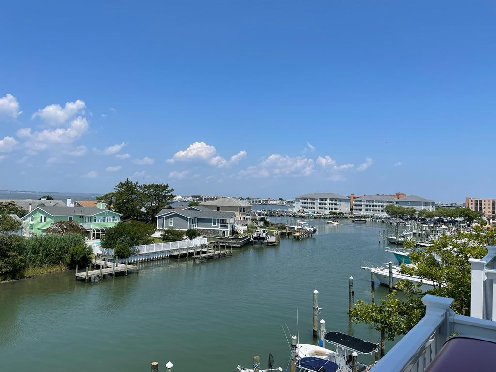 A view of a serene waterway is captured, with various boats docked along the banks. Charming homes in soft colors line the waterfront, and the sky is clear with a few scattered clouds, creating a tranquil atmosphere.