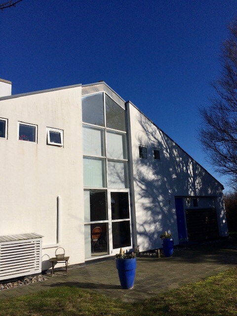 The exterior of the house is presented, featuring a modern design with large windows that enhance natural light. A well-maintained patio area shows potted plants in blue containers. A stone pathway leads to the entrance, and a glimpse of the surrounding greenery is visible.
