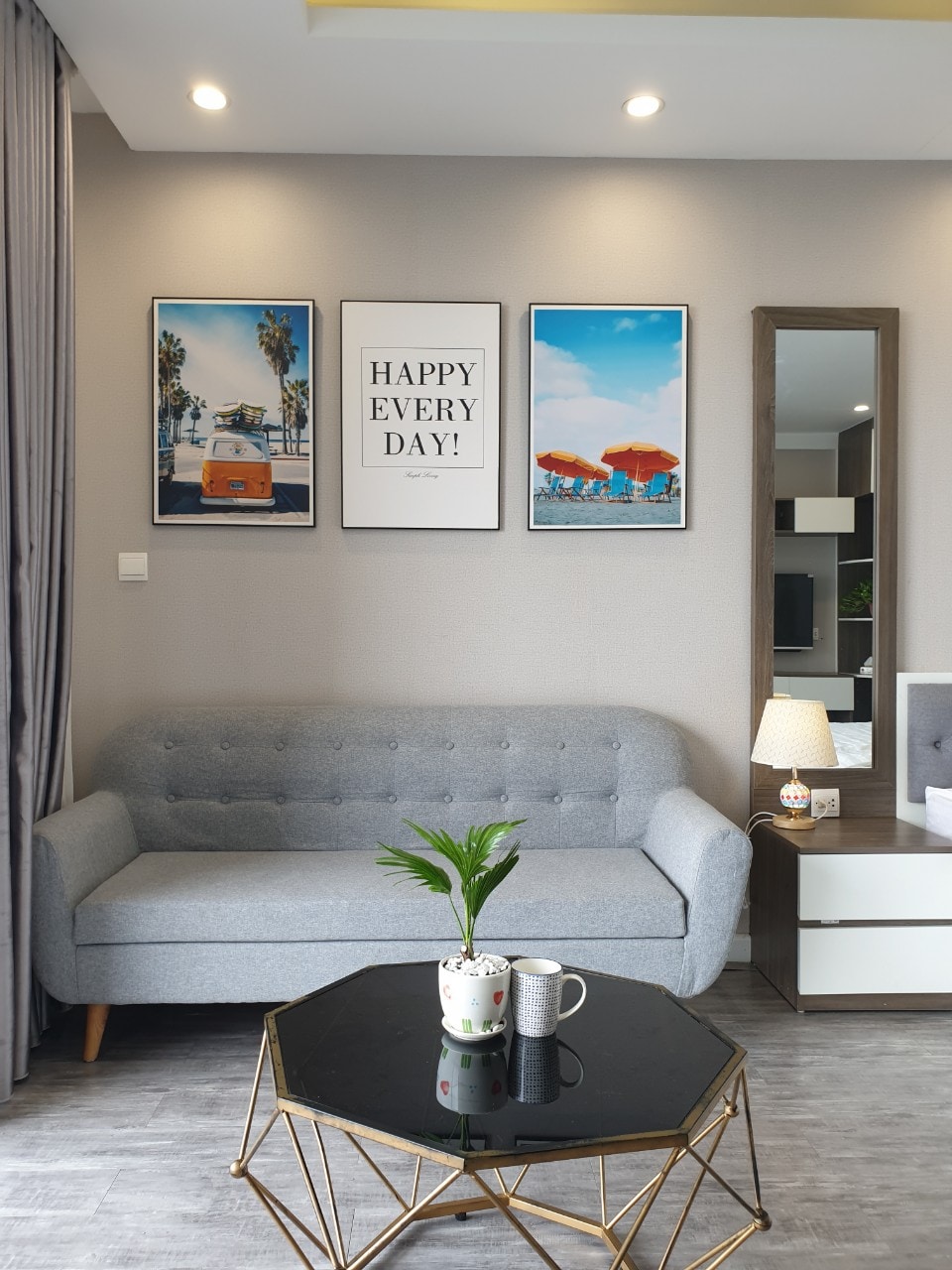 A modern living area features a light gray sofa complemented by a decorative coffee table with a geometric design. Three vibrant framed prints adorn the wall, and a mirror reflects the space. A small plant adds a touch of greenery to the coffee table.