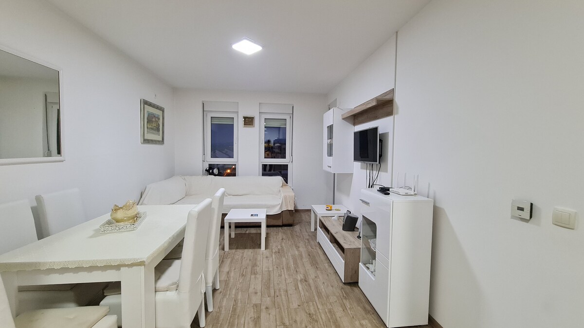 A modern living area features a light-colored sofa and a simple dining table with several chairs. A television is mounted on the wall opposite a white entertainment unit. Large windows allow natural light to fill the space, enhancing its inviting atmosphere.